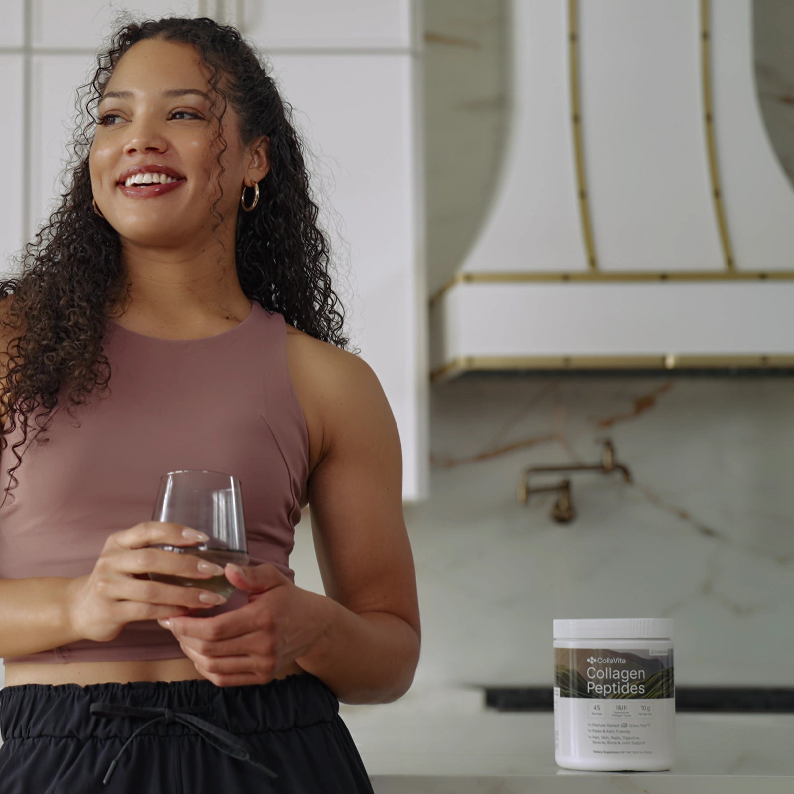 Smiling woman in activewear holding a glass of water next to a jar of CollaVita Collagen Peptides in a modern kitchen setting.